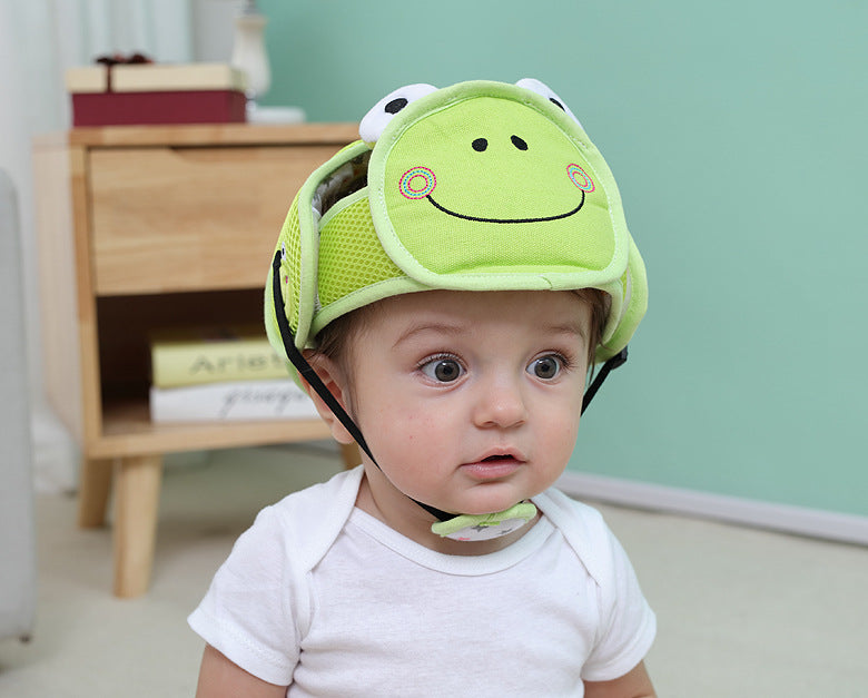 A baby wearing a green helmet with a smiling frog face design, sitting indoors with a wooden cabinet and books in the background. The baby is looking slightly surprised.