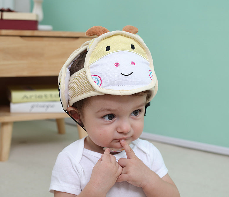 A toddler wearing an animal-themed protective helmet touches their lips with a finger while sitting indoors. The background has a wooden table and a mint green wall.