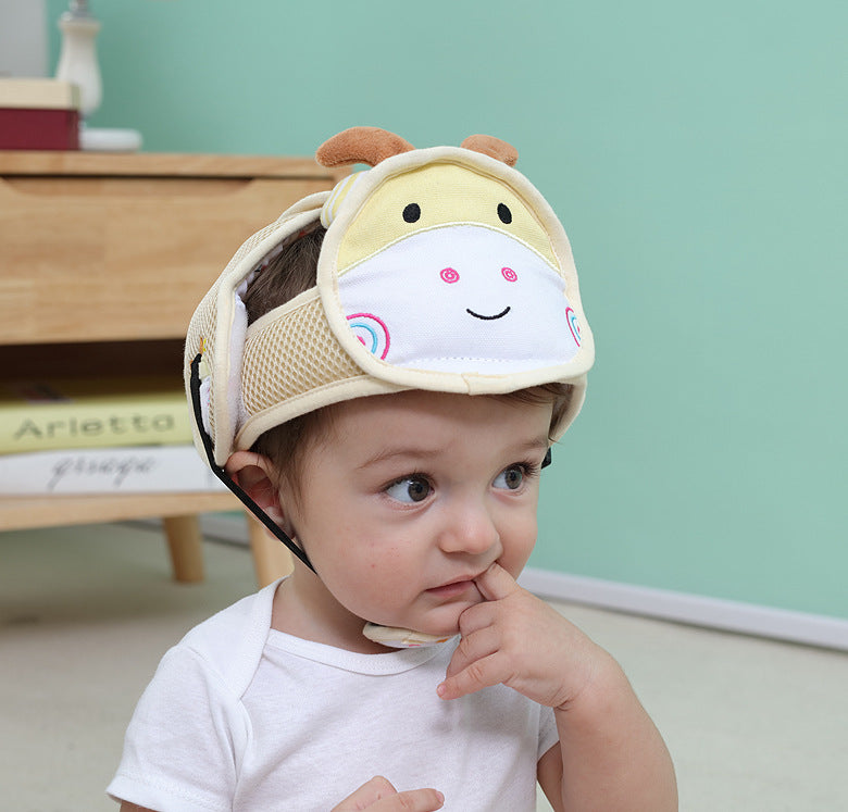 A baby wearing a light-colored helmet with a smiling animal face design, sitting indoors with a thoughtful expression and finger near the mouth, against a mint green wall background.