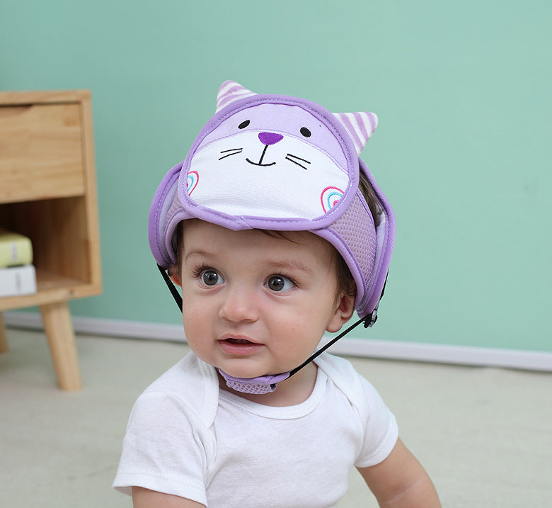 A baby wearing a white onesie and a purple cat-themed protective helmet with ears sits on the floor against a teal wall. A wooden shelf is partially visible in the background.