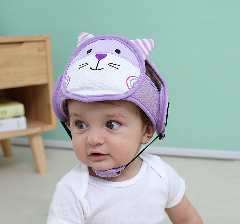 Baby wearing a cute purple helmet with a cat face design, sitting on the floor. In the background, there's a wooden shelf with books and a light green wall.