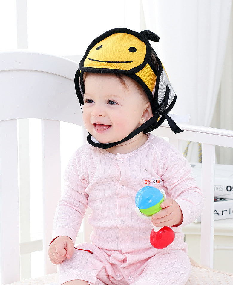 A smiling baby in a pink onesie sits on a white crib holding a colorful toy. The baby wears a yellow and black bee-themed protective headgear.
