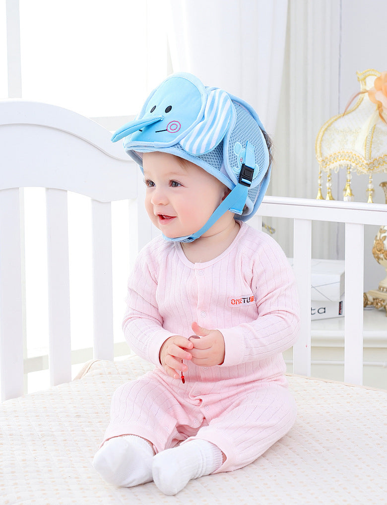 Cute baby wearing a blue elephant safety helmet sits on a crib, dressed in a pink onesie, against a soft, bright nursery background with a decorative lamp.