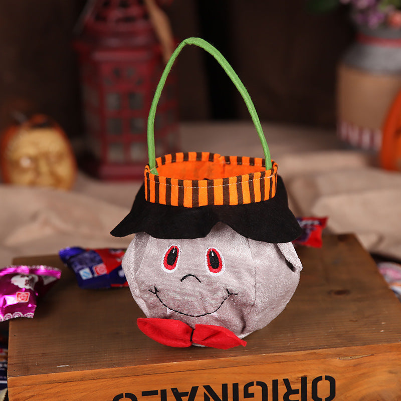 Plush Halloween basket with a smiling face, red bow tie, striped orange and black top, and green handle, placed on a wooden box with candy and decorative items nearby.