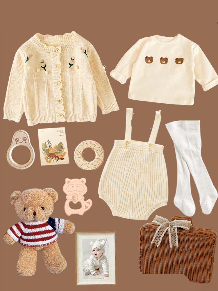 Baby outfit set on brown background, includes a cream knit cardigan, bear motif shirt, suspenders, tights, teddy bear, teething rings, framed photo, and a wicker basket.