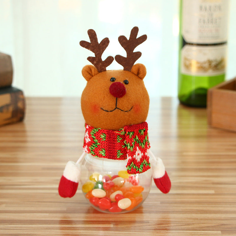 A festive reindeer decoration with antlers and a red nose, wearing a colorful knitted scarf, holds a clear jar filled with assorted jelly beans on a wooden surface.