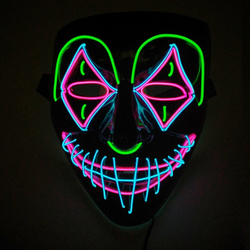 A neon mask with vivid designs: green brows, pink triangles around eyes, and blue and pink stitched smile, glowing against a dark background.