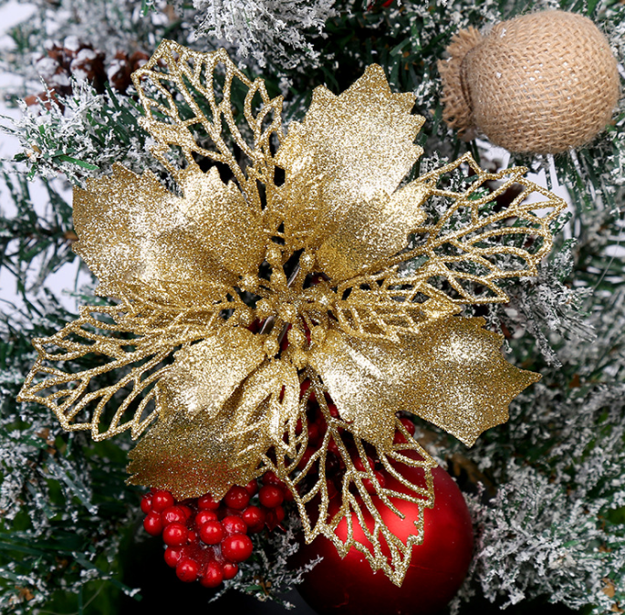 Golden glittery poinsettia decoration with red berries and ornaments on a snow-frosted Christmas tree branch, accented by a small burlap-wrapped decoration above.