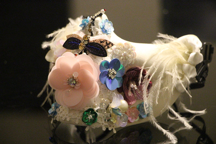 A decorative face mask adorned with pink and blue flower embellishments, feathers, and shimmering rhinestones, featuring a butterfly detail, on a reflective surface.