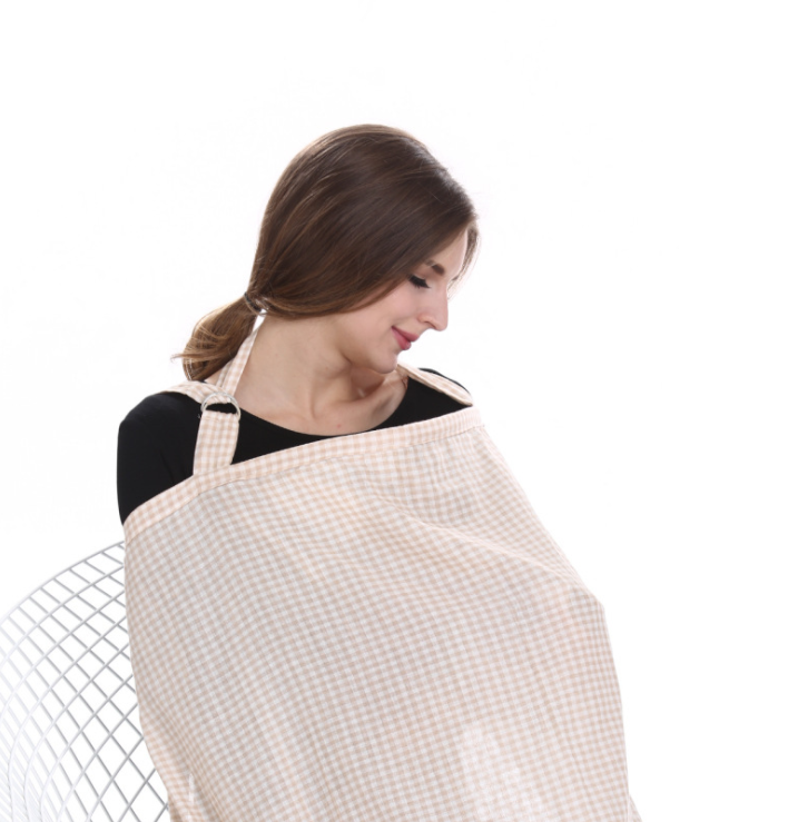 A woman sits on a white chair, wearing a black top and a gingham-check nursing cover, looking down with a gentle smile. The background is plain white.
