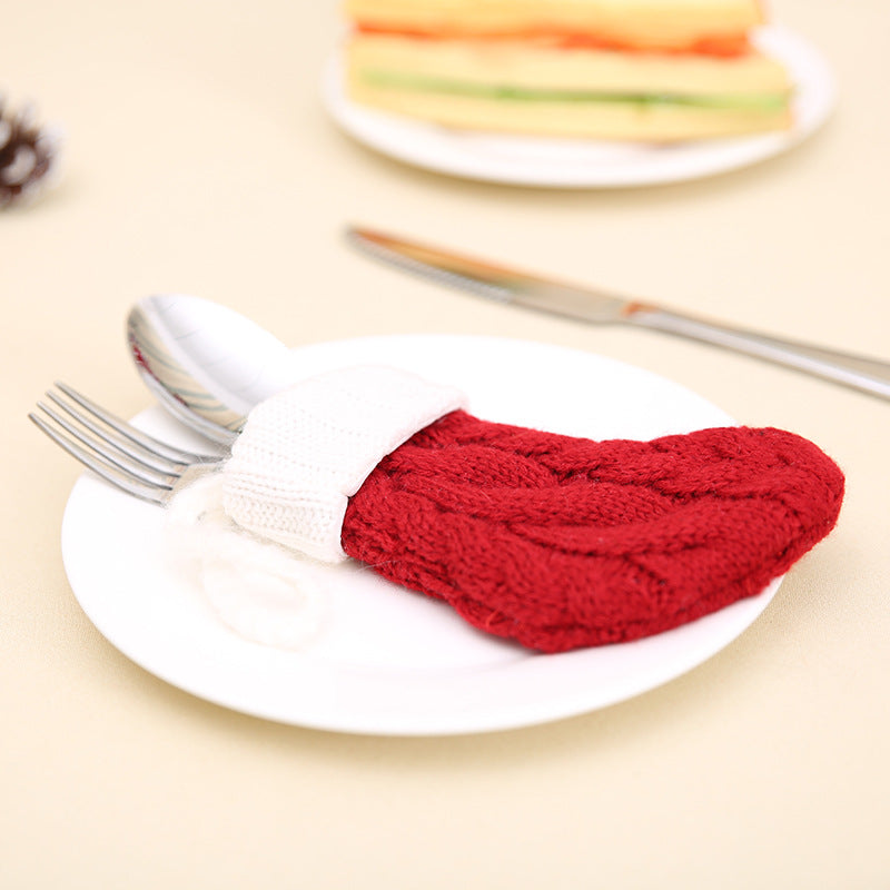 Red knitted stocking holds silverware on a white plate. A knife lies beside it and a blurred plate of sandwiches is in the background.