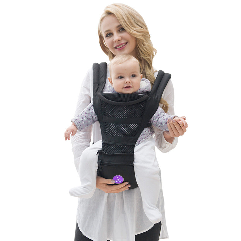 A woman smiling while carrying a baby in a black mesh front baby carrier. The baby wears a patterned shirt and appears content, with both holding hands.