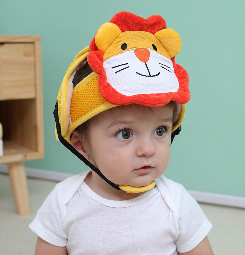A baby wearing a lion-themed protective helmet sits attentively. The helmet is yellow with a red mane and a lion face on the front. The background is a soft green wall.