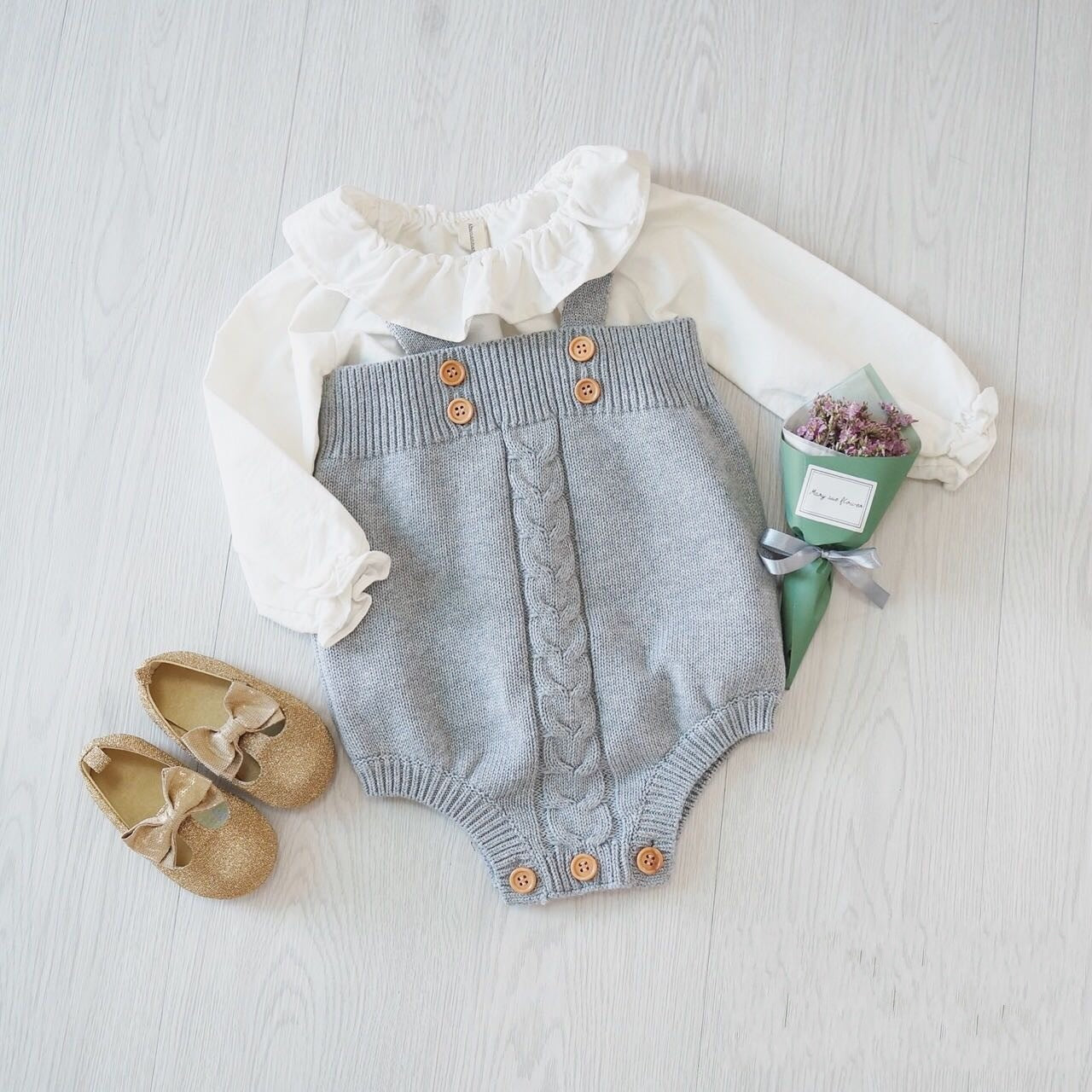 Baby outfit with a white ruffled blouse, grey knit romper with buttons, and gold shoes with bows, laid on a light wood floor. A small bouquet of purple flowers is placed nearby.