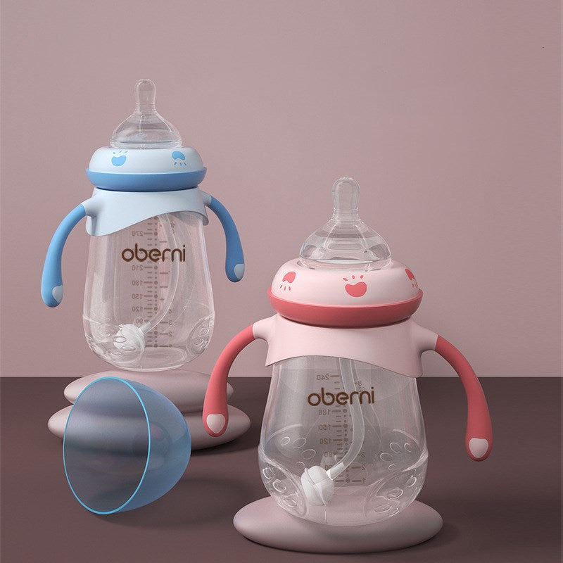Two baby bottles with handles sit on a pink surface. One is pink, and the other is blue. Both have clear bodies, measurement markings, and are labeled "Oberni."