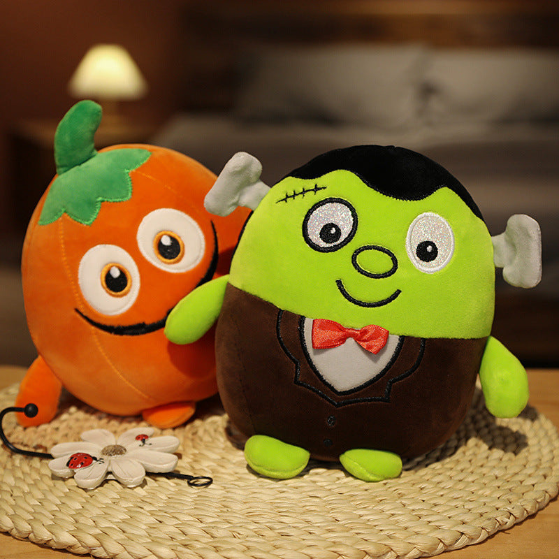Two plush toys, a green Frankenstein with a red bow tie and a smiling orange pumpkin, sit on a woven mat. A small flower decoration is next to them.