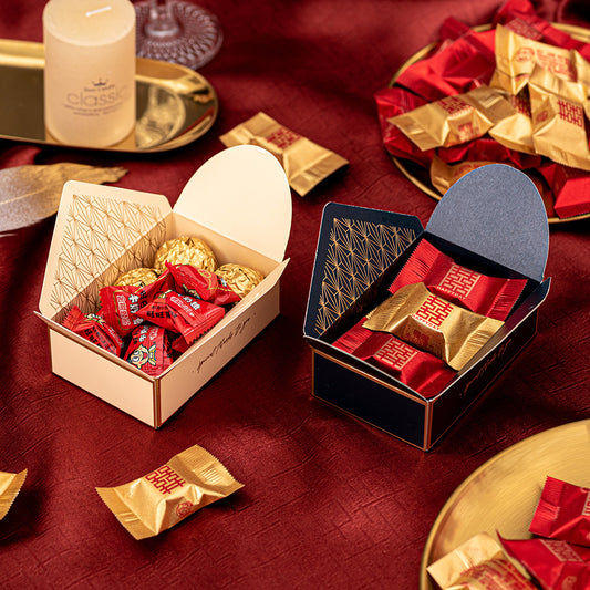 Two open gift boxes on a red cloth, one with gold-wrapped chocolates and red candies, the other filled with red and gold-wrapped chocolates. A candle and more candies are on the table.