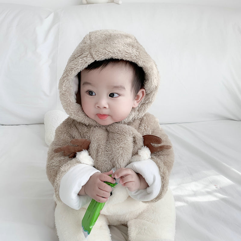 A baby in a fluffy brown onesie with antler details sits on a white couch, holding a green item, looking to the side with a curious expression.