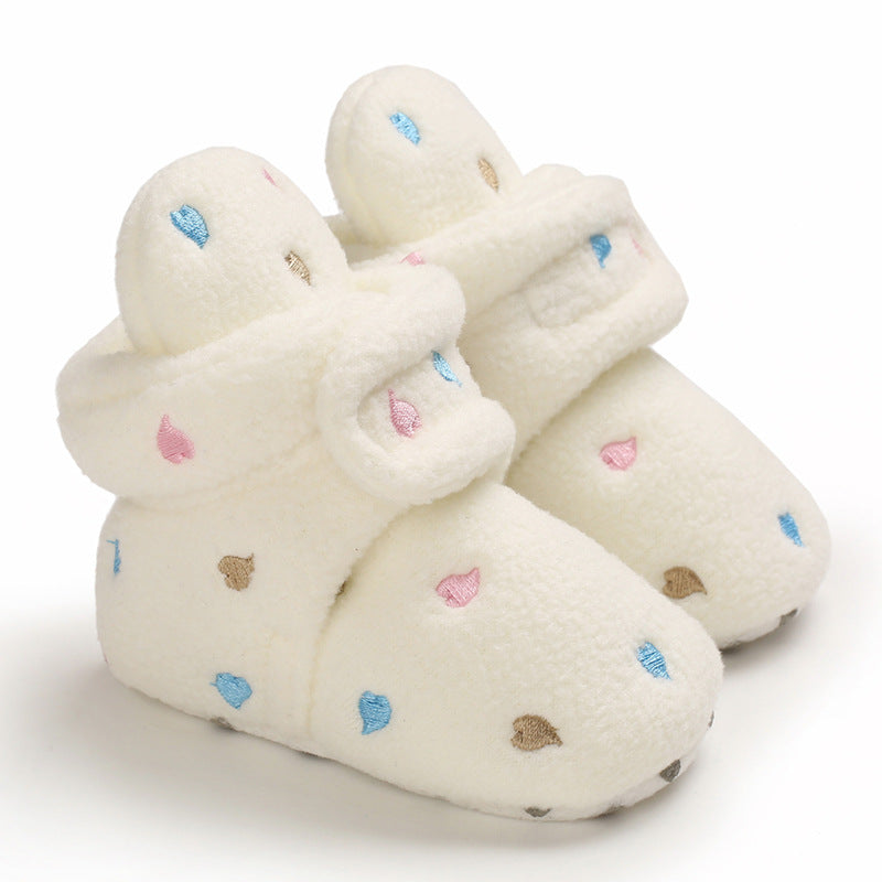 Cozy white baby booties with soft plush texture, adorned with small blue, pink, and brown embroidered raindrop patterns, and Velcro strap closure, set against a plain background.