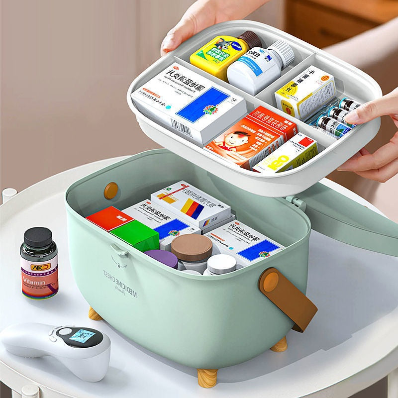 Mint green medicine box on a table holds various medicine bottles and boxes, with a transparent organizer tray above. Nearby, a vitamin bottle and digital thermometer are visible.