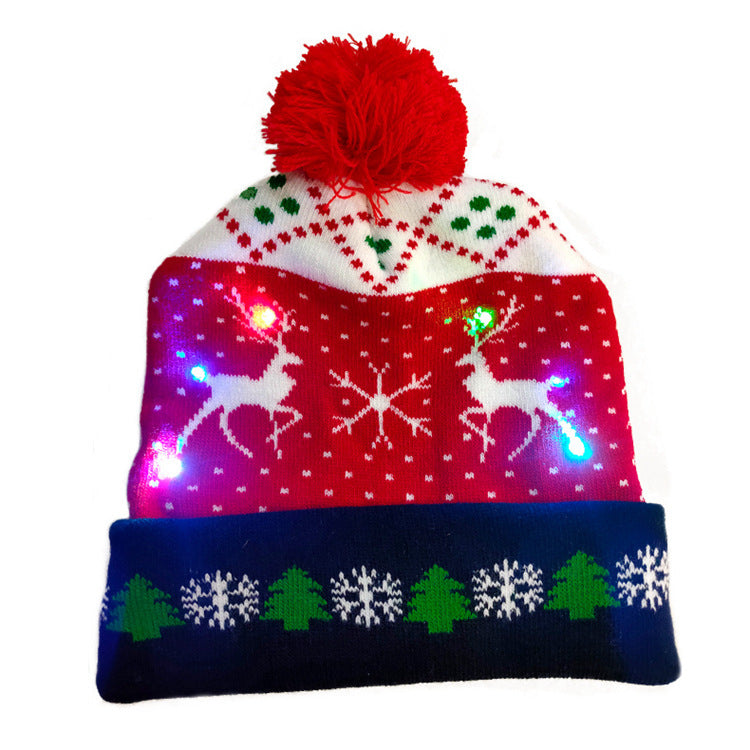 Festive knit hat with a red pom-pom, featuring reindeer, snowflakes, and green trees. The design includes white, red, and green patterns with colorful LED lights.