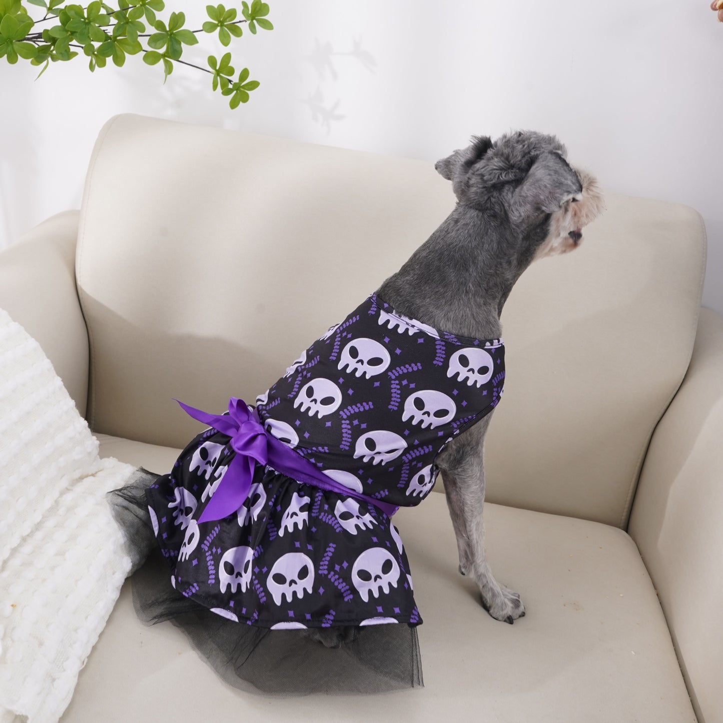 A gray dog on a beige couch wears a purple dress with skull patterns and a bow. Lush green leaves hang above, casting soft shadows on the wall.