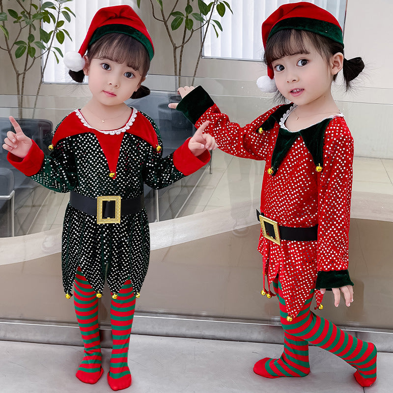 Two young children in festive elf costumes with red hats stand indoors, smiling. One wears black with white polka dots; the other red with green accents and striped tights.
