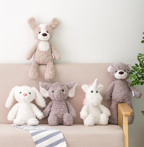 Five plush toys, including a dog, bunny, elephant, unicorn, and bear, are arranged on a beige couch. A striped blanket is draped over the armrest, and a plant is partially visible.