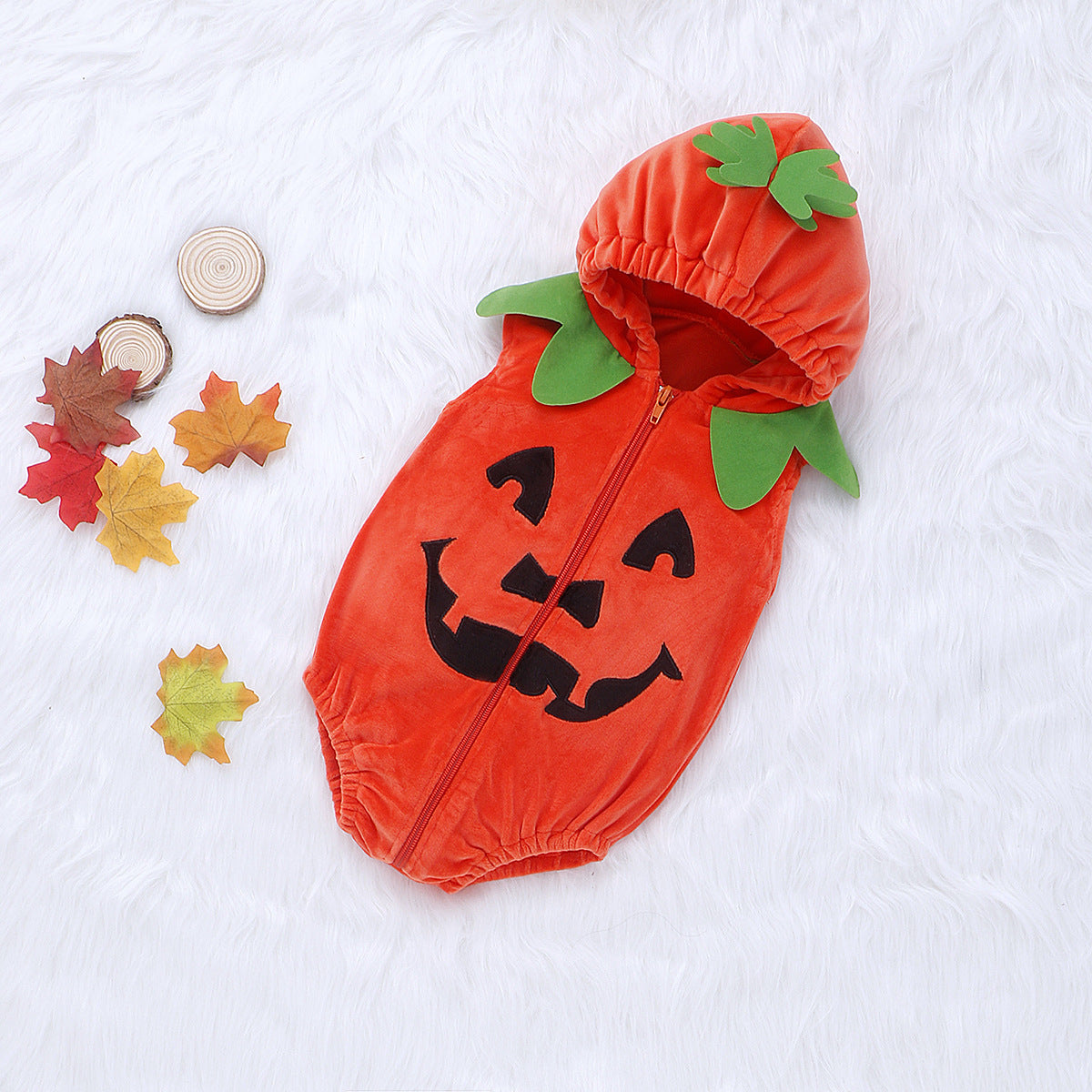 Orange pumpkin-themed baby romper with green leaf hood and smiling jack-o'-lantern face, on a white furry background, surrounded by autumn leaves and wooden discs.