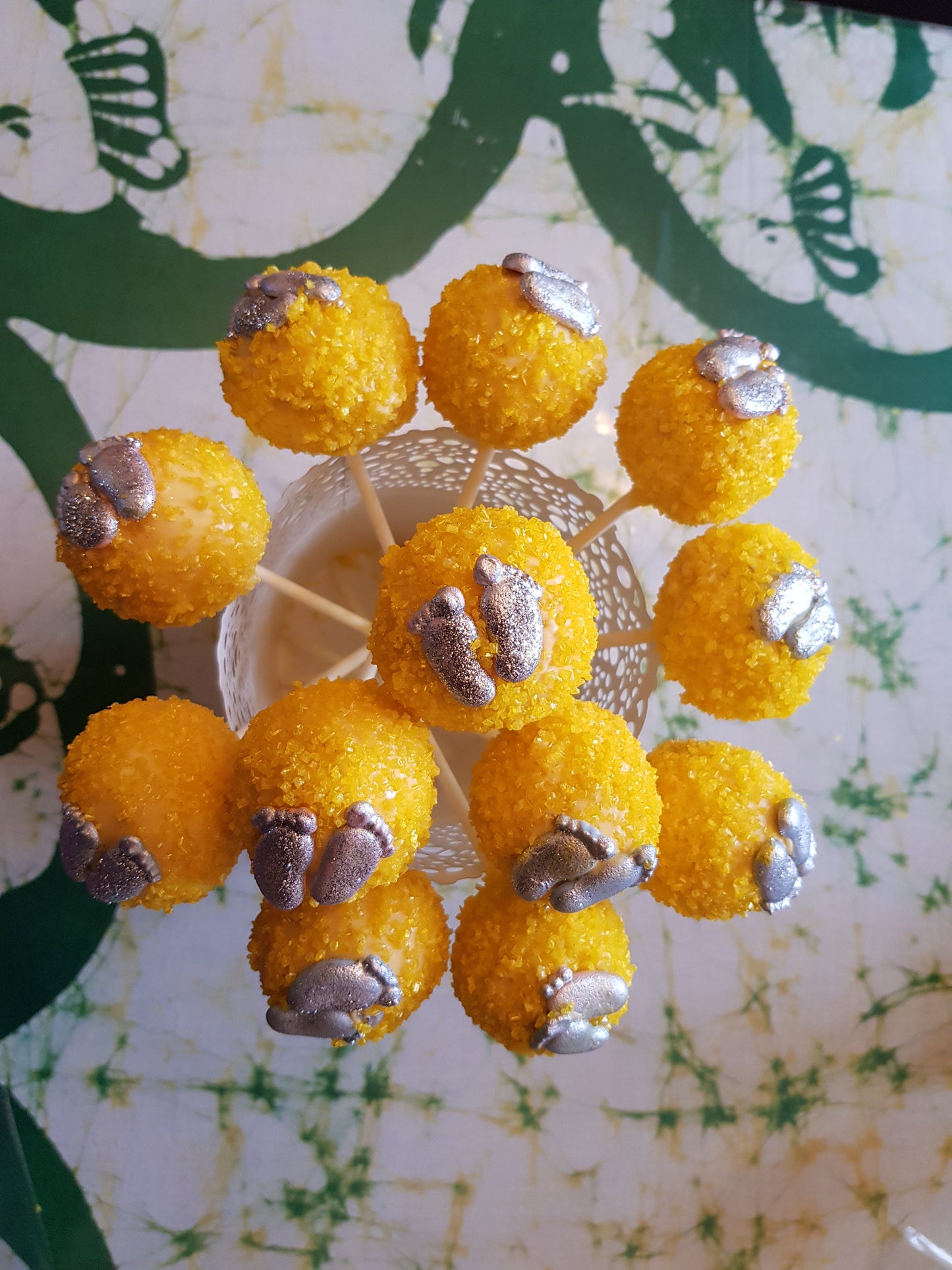 Yellow cake pops with silver foot decorations, arranged in a circular holder on a green-and-white patterned surface.