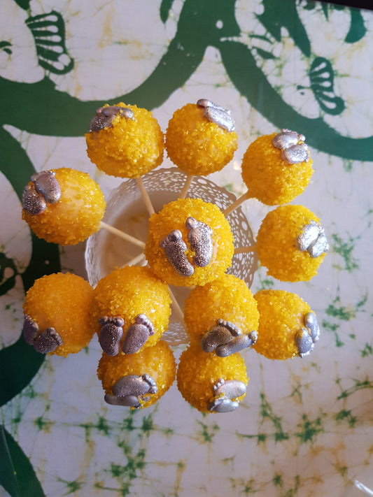 Yellow cake pops with silver foot decorations, arranged in a circular holder on a green-and-white patterned surface.