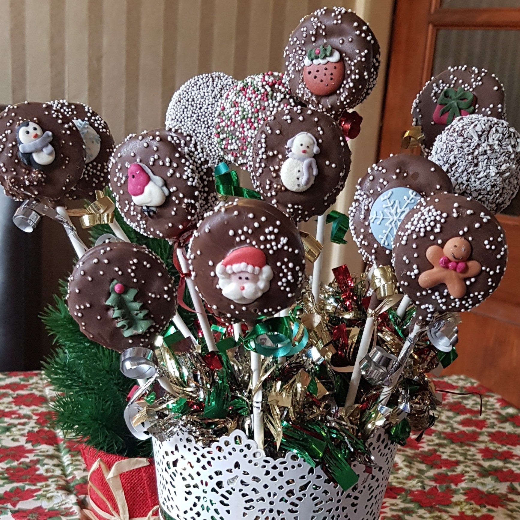 Festive chocolate pops with holiday motifs like Santa, snowmen, and snowflakes on sticks, arranged in a decorative pot with ribbons, set on a Christmas-themed tablecloth.