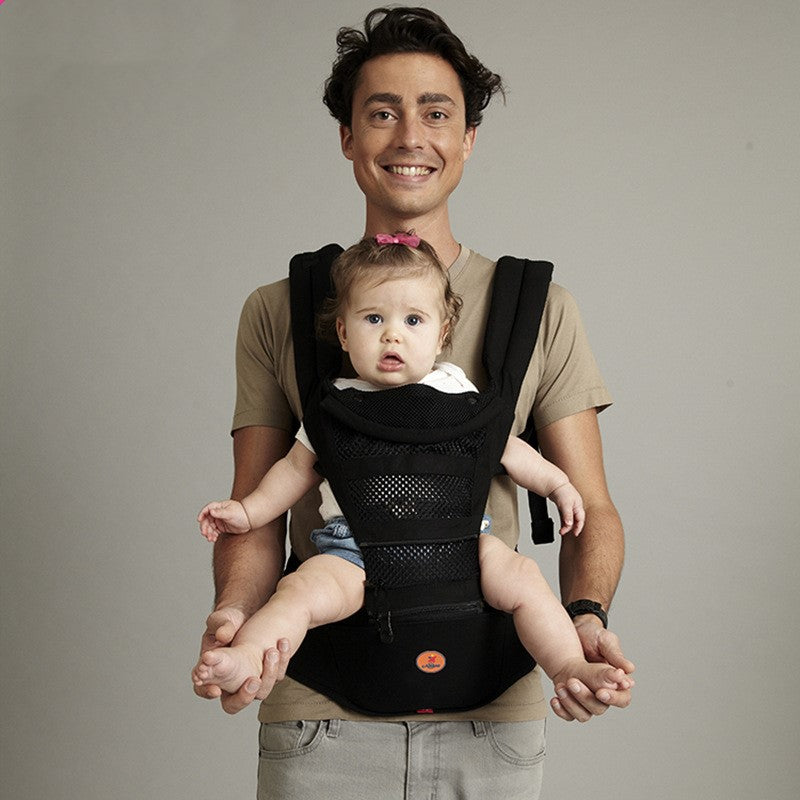Smiling man in a beige shirt holds a baby in a black baby carrier. The baby, wearing a white outfit and pink bow, looks forward with a curious expression against a gray background.