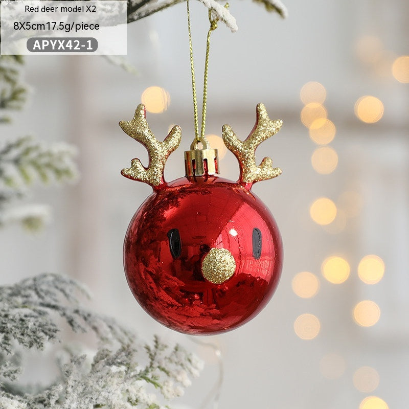 Red Christmas ornament shaped like a reindeer's face with glittery gold antlers and nose, hanging on a snow-dusted tree branch with blurred warm lights in the background.