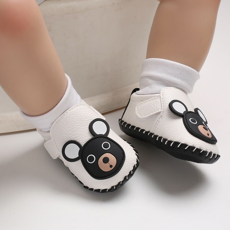 Baby wearing white shoes with cute bear face patch, white socks, and sitting on a light-colored surface. The shoes have a textured surface and black stitching.