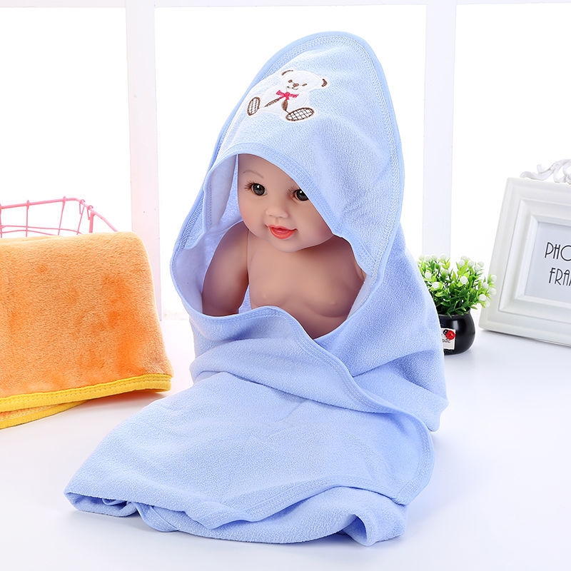 A baby doll wrapped in a light blue towel with a bear design, sitting on a white surface. Nearby are a folded orange towel, a small plant, and a photo frame.