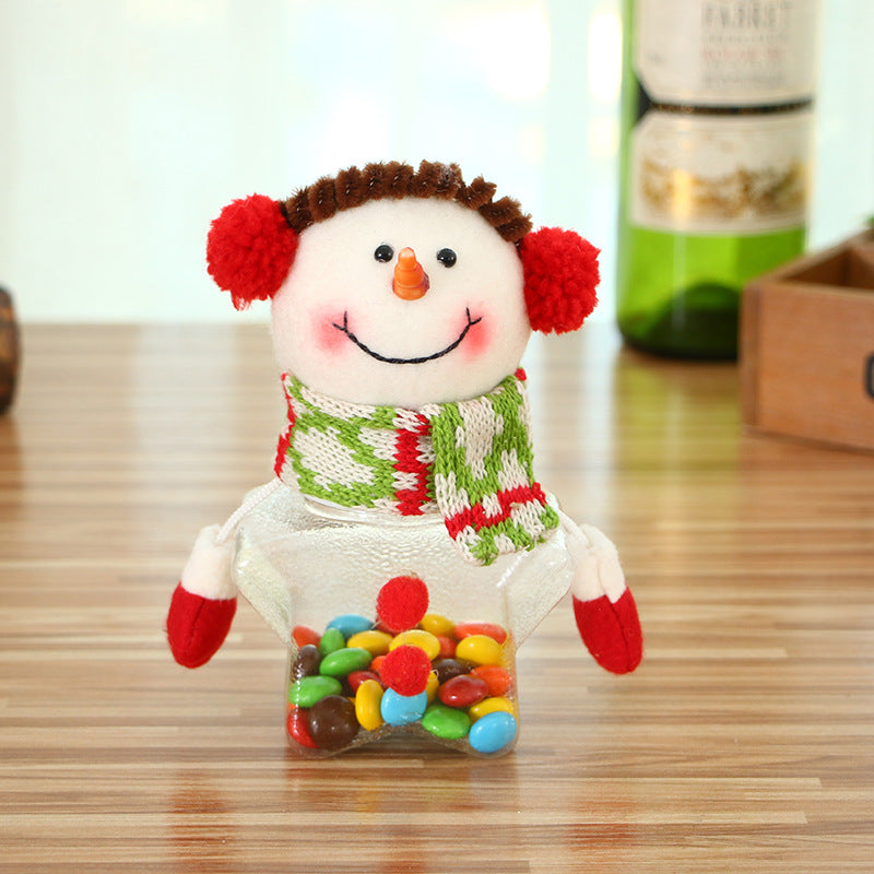 A festive snowman container with red earmuffs, carrot nose, and colorful scarf sits on a wooden table. It holds colorful candy in its star-shaped base.