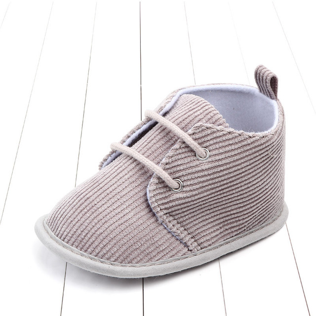 A soft, beige baby shoe with corduroy texture, white sole, and gray laces. The shoe is on a white wooden floor, viewed from a slight angle.