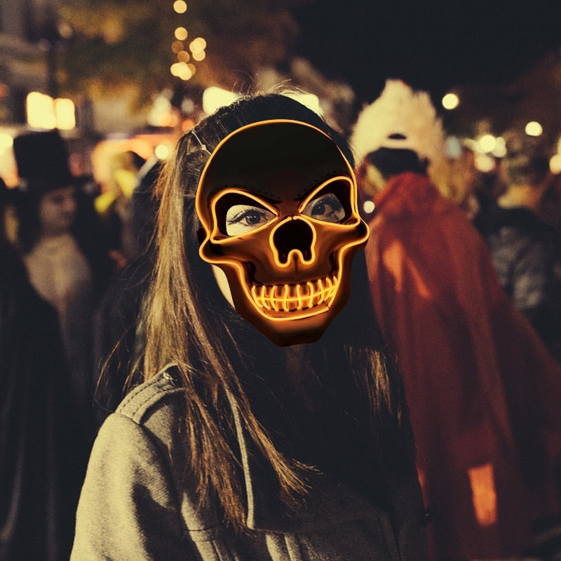 Person wearing a glowing skull mask stands in a festive, crowded outdoor setting at night, with blurred lights and people in costumes in the background.