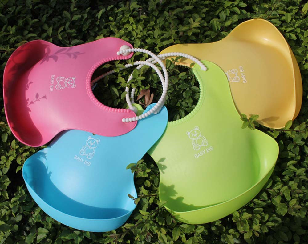 Four colorful silicone baby bibs in pink, yellow, green, and blue are laid out on green foliage. Each bib has a teddy bear design and beaded straps.