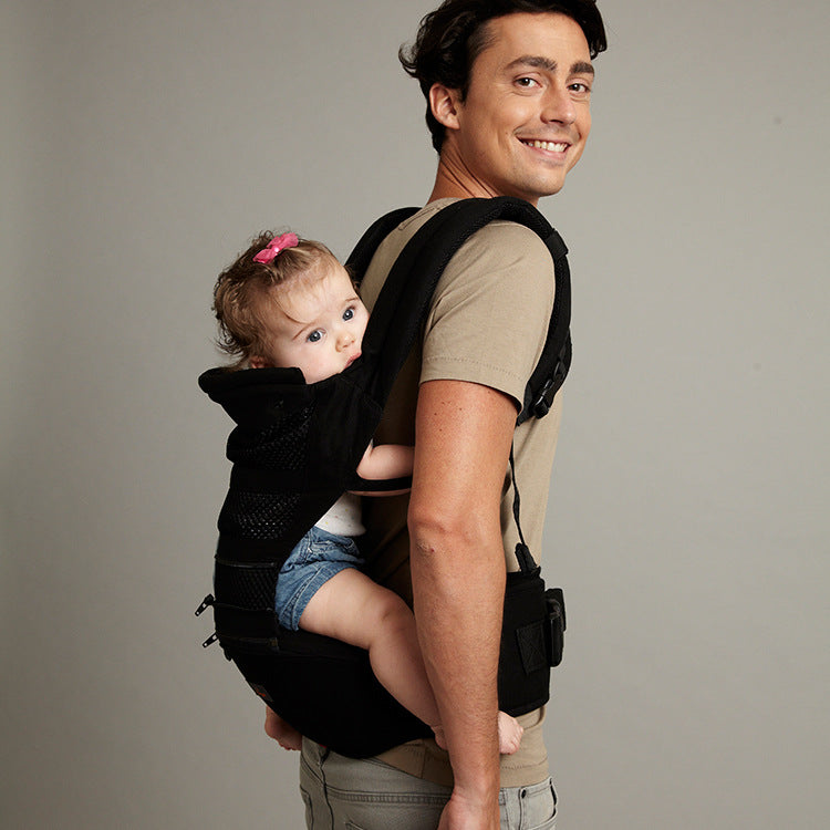 Smiling man wearing a beige shirt carries a baby in a black carrier on his back. The baby, with a pink bow in her hair, looks to the side.