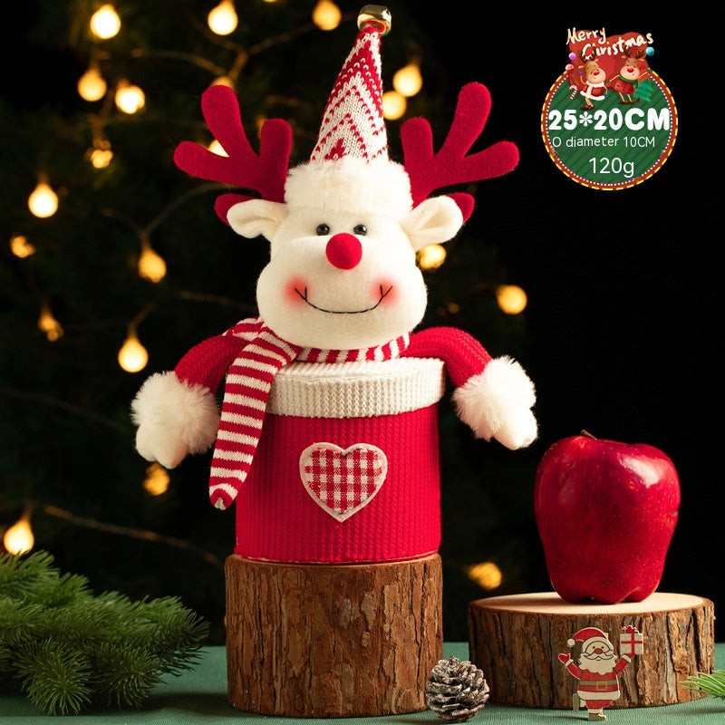 Plush reindeer in a red outfit with antlers, scarf, and heart patch, sits on a log. An apple and Santa figurine are beside it. Background features a lit Christmas tree.