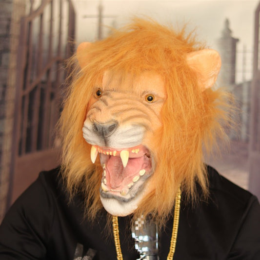 Person wearing a realistic lion mask with an open mouth, detailed mane, and long teeth. The mask is paired with a black outfit and gold chain, set against a blurred backdrop of gates.
