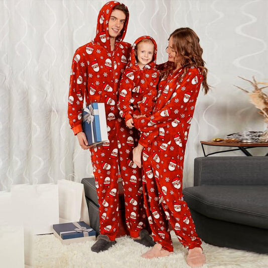 Family wearing matching red holiday pajamas with Santa and snowflake prints. The young child is held by the mother while the father holds a gift box. Cozy living room setting.