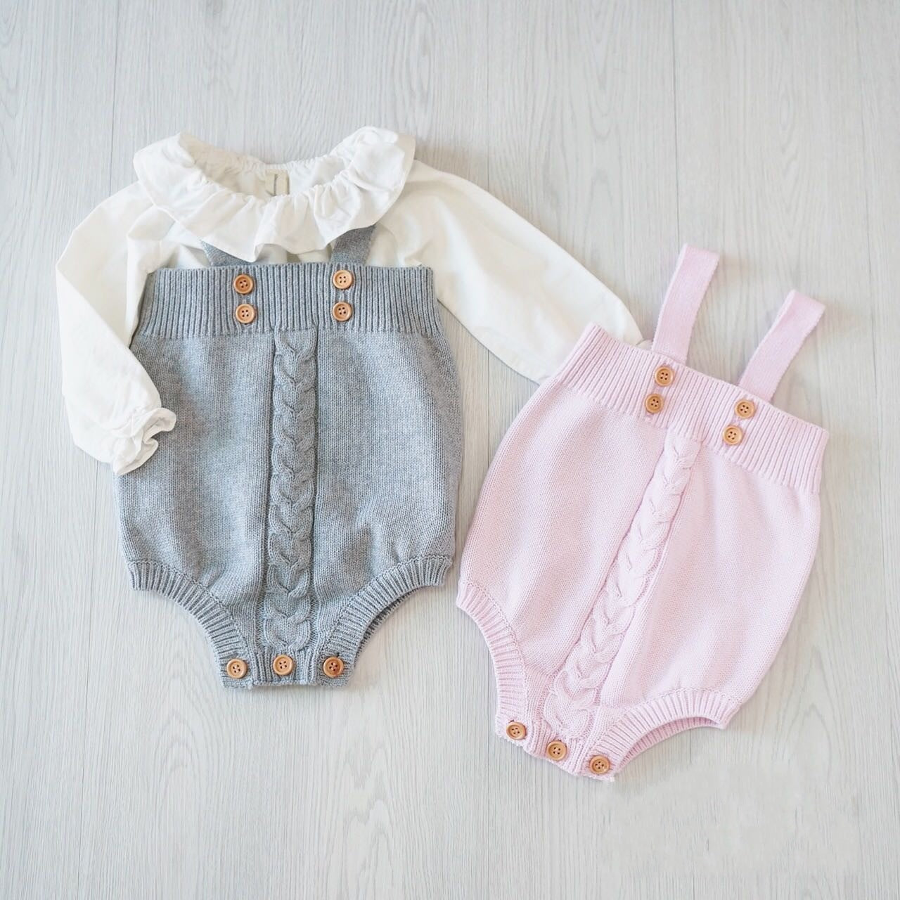 Two baby rompers on a light wooden floor: one gray with a white ruffled top, both with cable-knit patterns and wooden buttons, the other pink with suspenders.