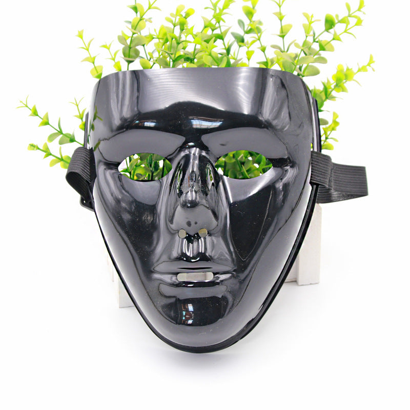 A glossy black face mask with cut-out eyes and mouth is set against a backdrop of small, bright green leaves. A black strap is attached on each side of the mask.
