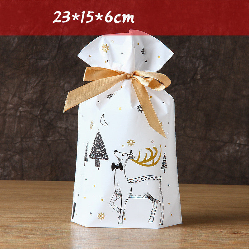 White gift bag with gold ribbon, featuring a reindeer and tree design with snowflakes. Dimensions are 23x15x6cm, displayed on a wooden surface against a dark background.