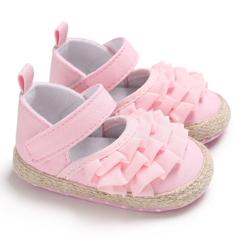 Pink baby shoes with frilly embellishments and Velcro straps, featuring a woven straw-like trim around the sole. Ideal for infants, these shoes offer a cute, comfortable design.
