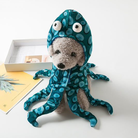 A teddy bear dressed in a teal octopus costume with blue spots and big eyes. It sits on a white surface near an open box and a yellow card with plant images.