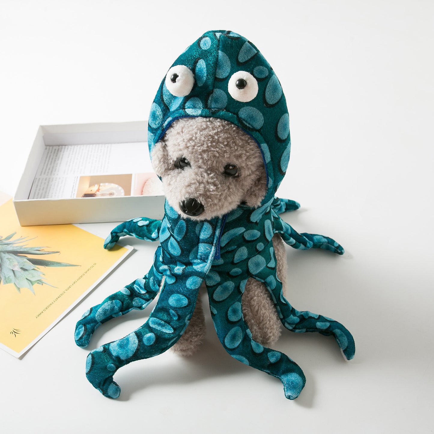 A gray plush dog wears a blue, polka-dotted octopus costume with large eyes. It sits on a table next to an open box and a yellow book with a plant image.